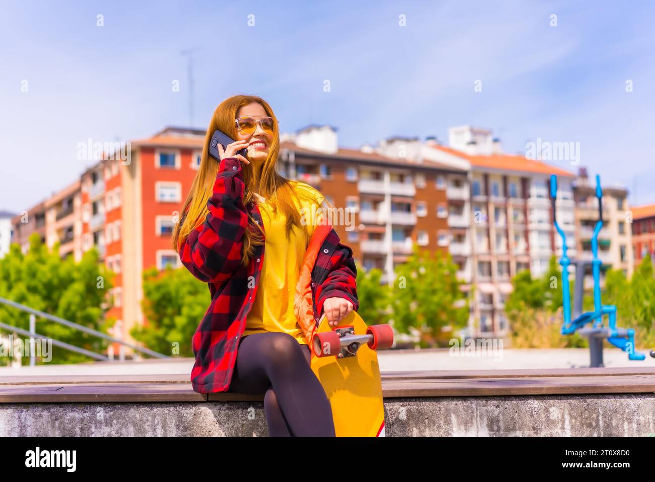 Skater woman in a yellow t-shirt, red plaid shirt and sunglasses, sitting with skateboard in the city, smiling calling with the phone Stock Photo