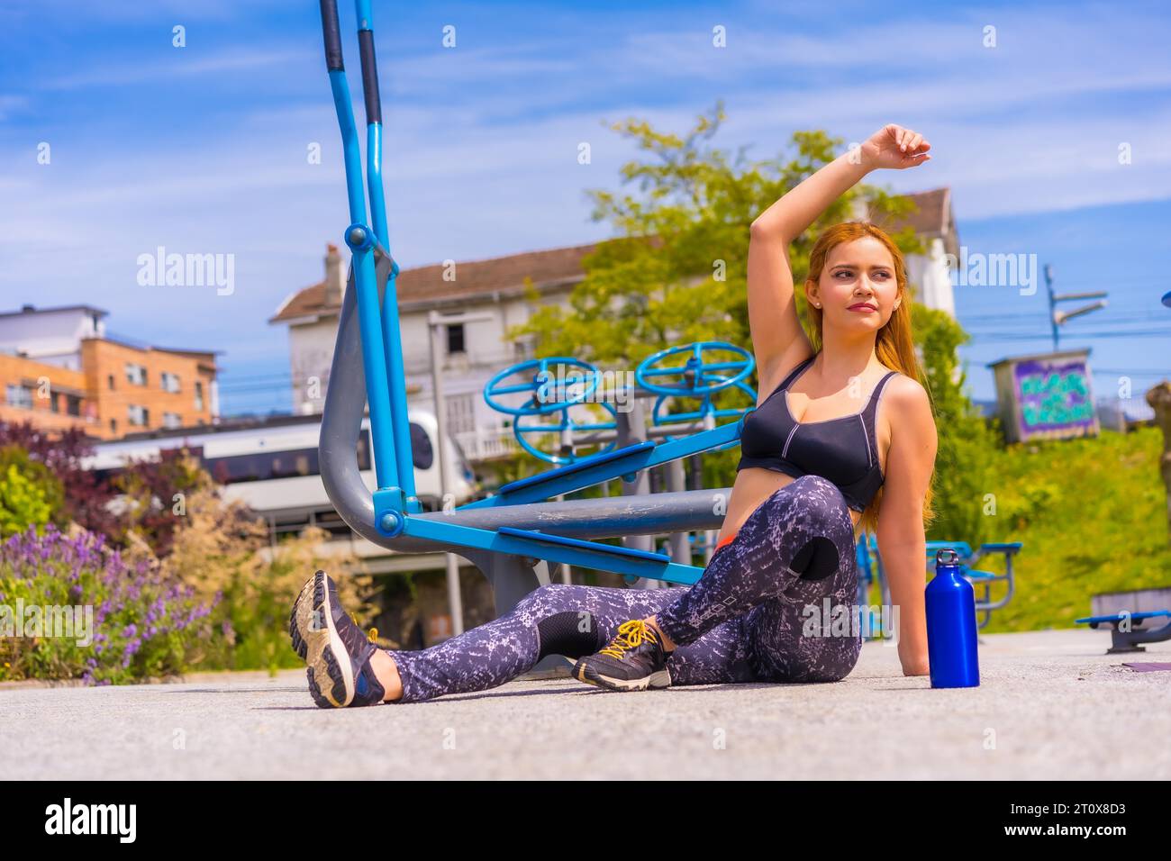 Red-haired woman with purple leggings and black overalls exercising on machines, stretching, sport in the city Stock Photo