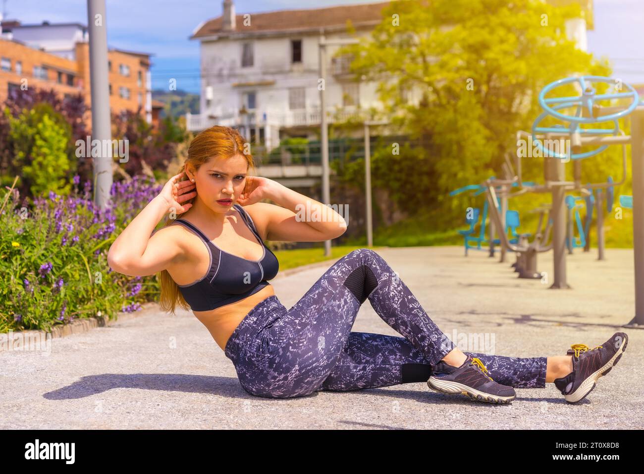 Red-haired woman with purple leggings and black overalls exercising on the machines, doing sit-ups, sport in the city Stock Photo