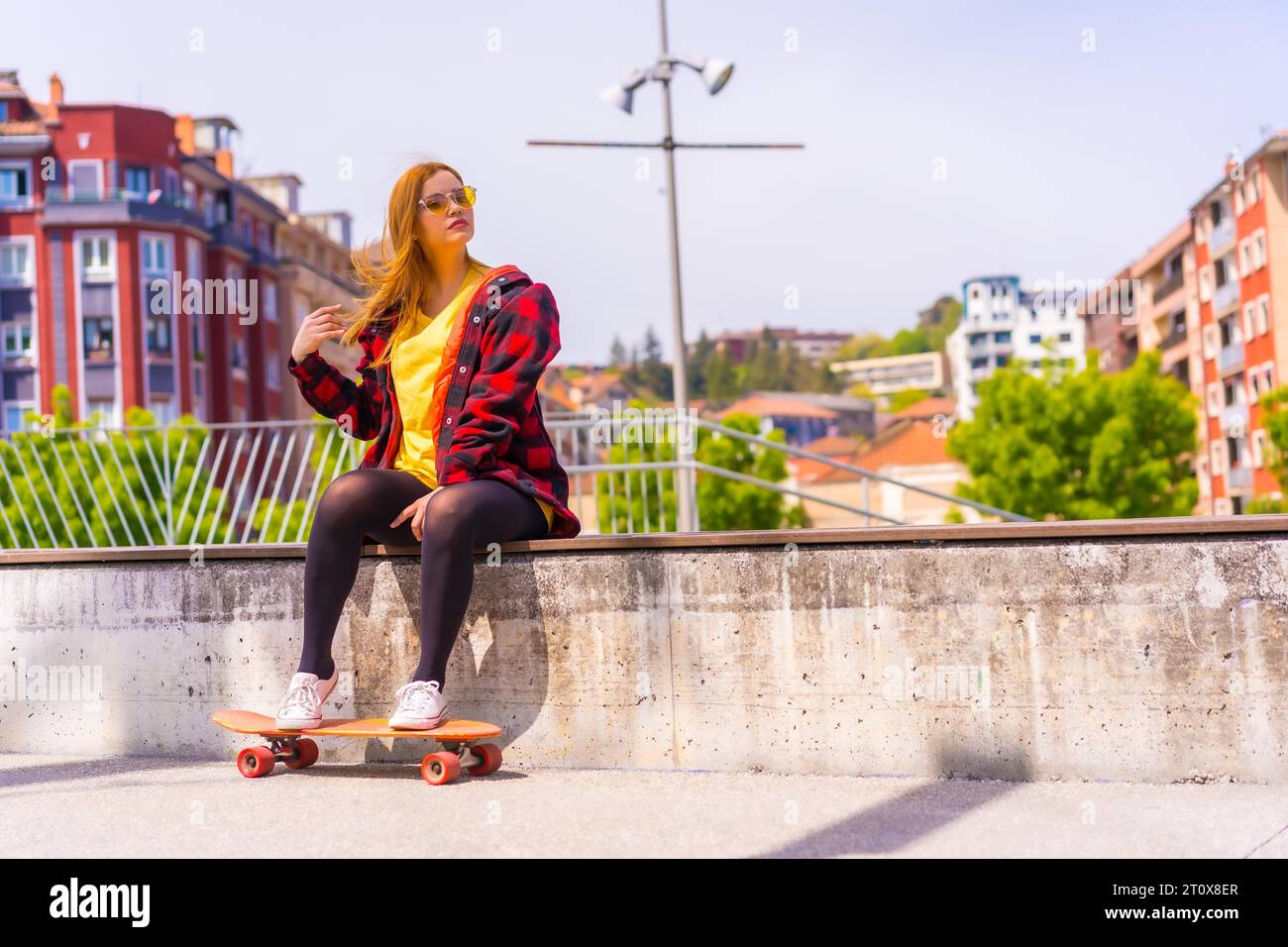 Skater woman in a yellow t-shirt, red plaid shirt and sunglasses, sitting with skateboard on a bench in the city Stock Photo