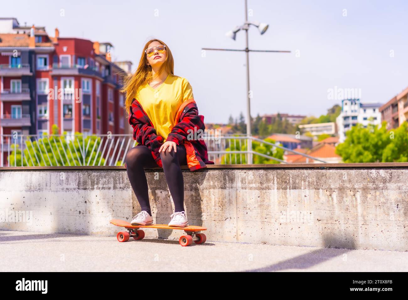 Skater woman in a yellow t-shirt, red plaid shirt and sunglasses, sitting with skateboard on a bench in the city Stock Photo