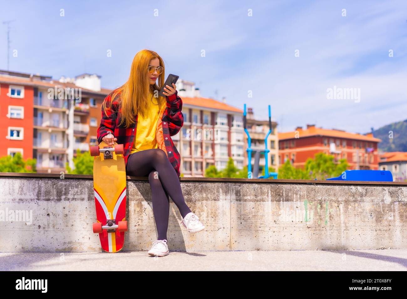 Skater woman in a yellow t-shirt, red plaid shirt and sunglasses, sitting with skateboarding in the city sending an audio message with the phone Stock Photo