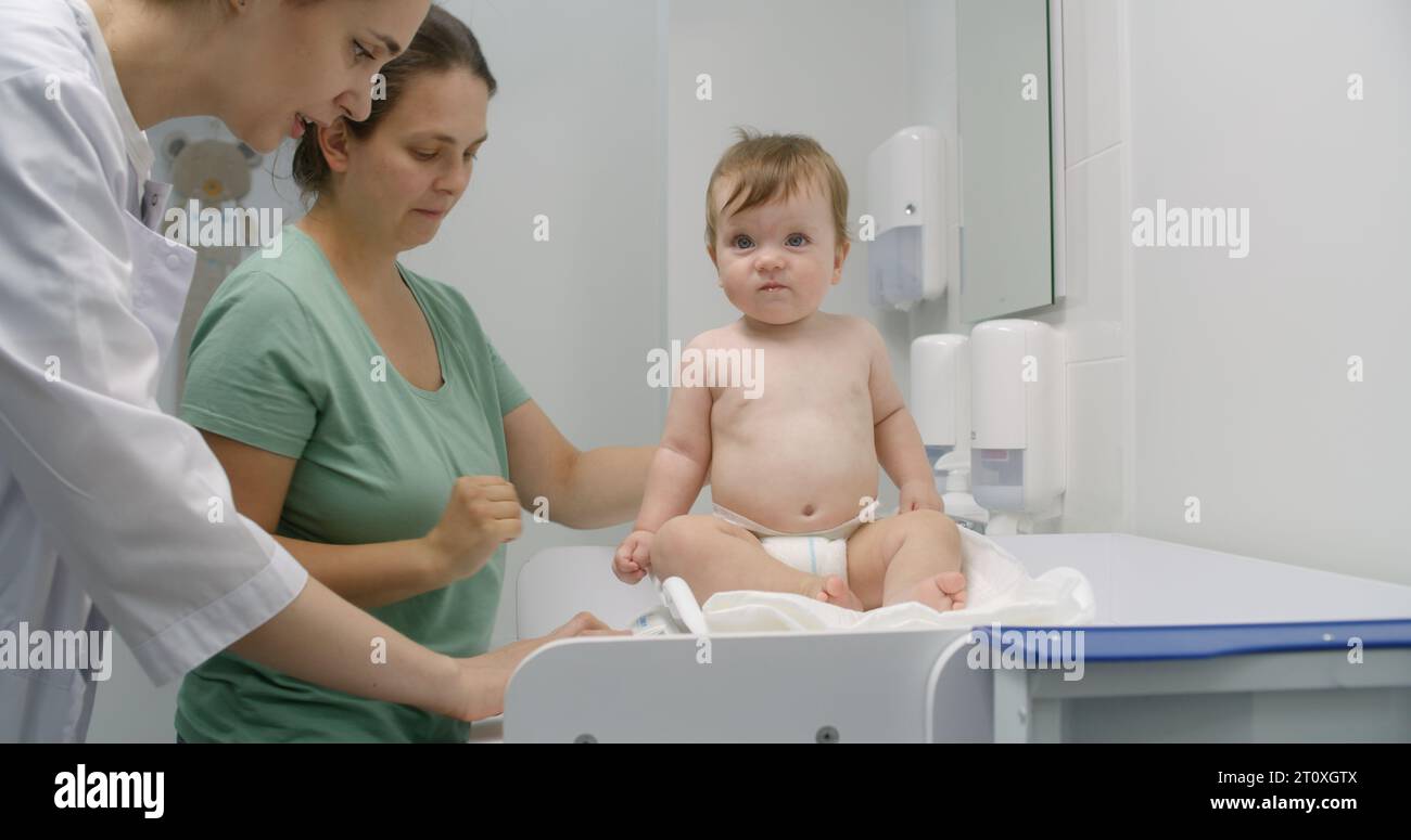 Mother holds baby on hands. Medic puts scale on changing table. Female ...