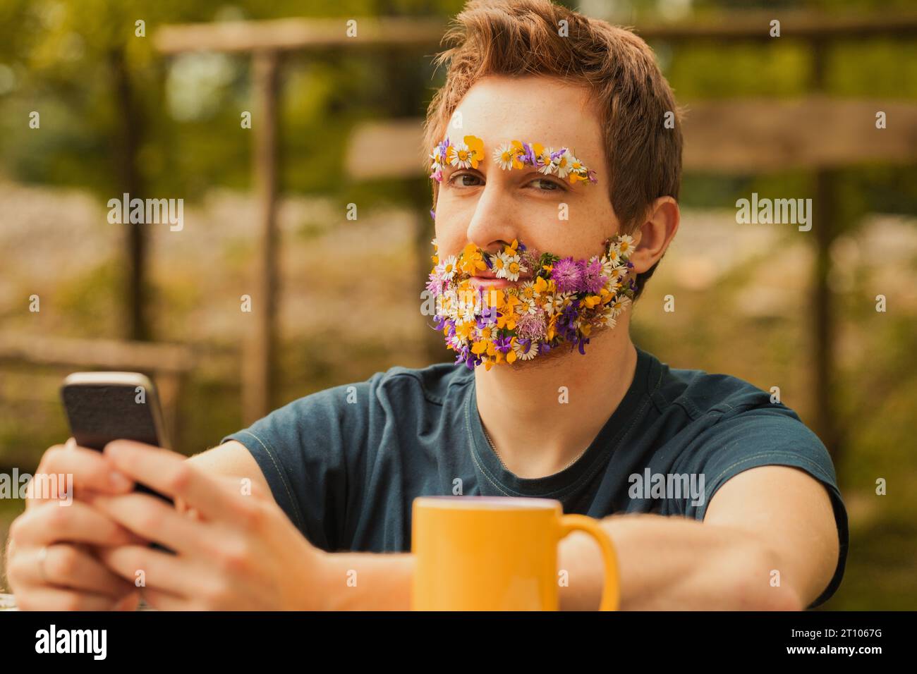 Young man with colorful spring flowers instead of facial hair sits amidst nature. While cherishing the tranquility and fresh air, he doesn't despise t Stock Photo