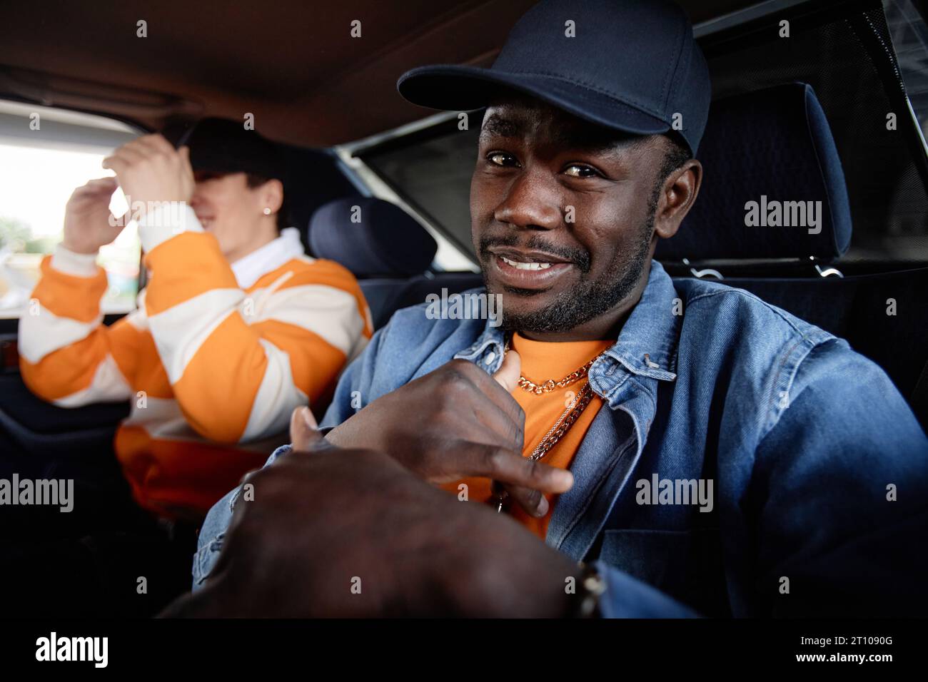 Young smiling African American rapper in cap and denim jacket looking at camera while sitting on backseat of car against his buddy Stock Photo