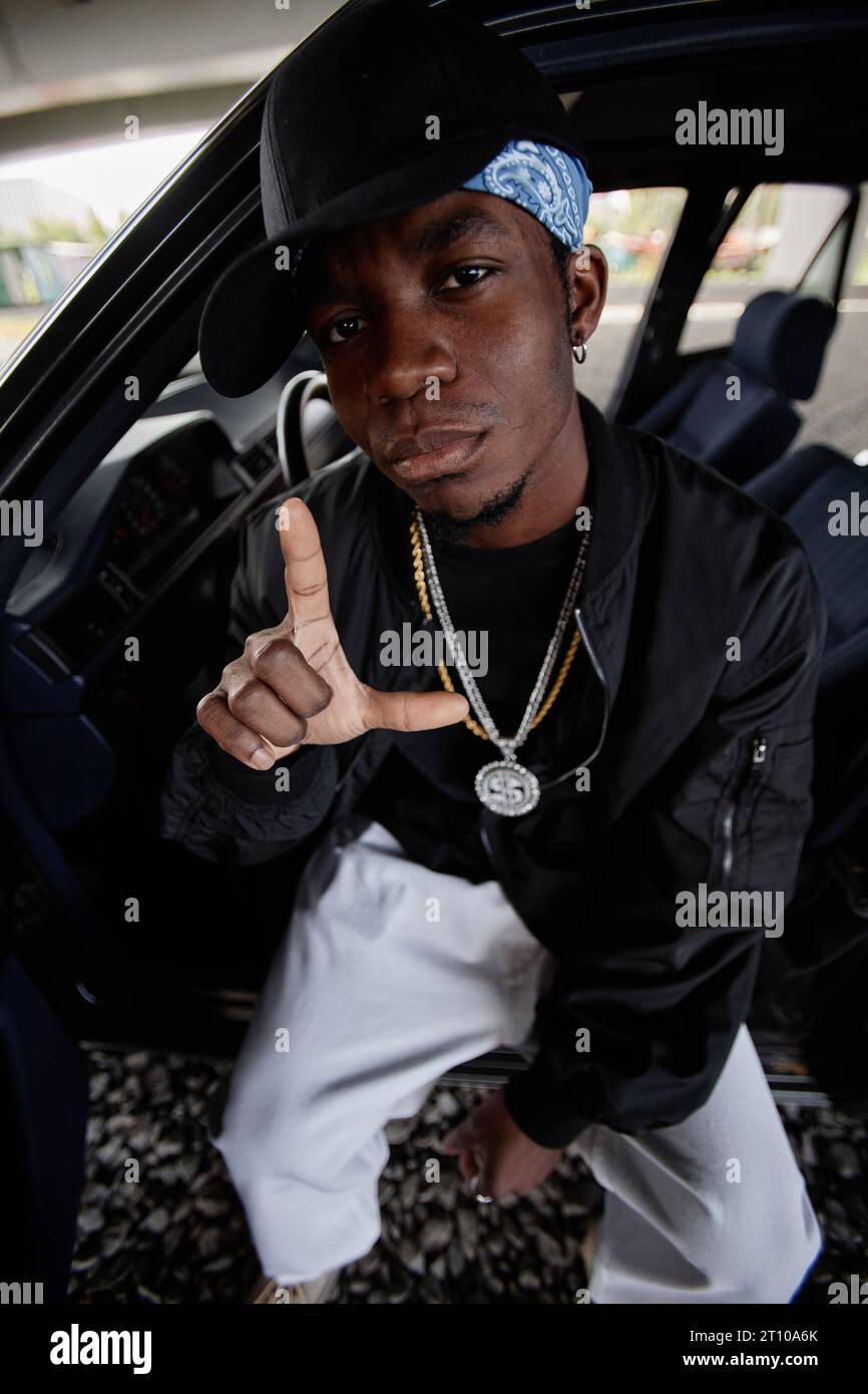 African American hip hop dancer in black bomber jacket, cap and white jeans sitting by open door in front seat of car and looking at camera Stock Photo