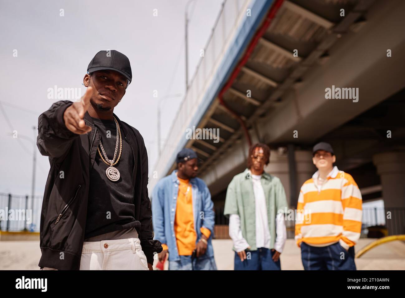 Young African American man in black cap and bomber jacket pointing at you while standing against group of intercultural buddies outdoors Stock Photo