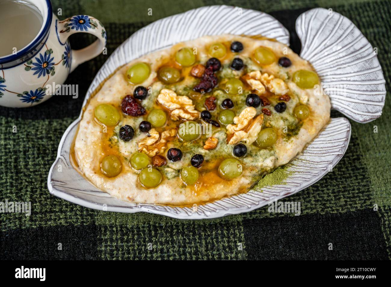 Porridge oatmeal with fruit, nut, honey and moringa powder in decorative fish-shaped plate on green checkered green wool tablecloth, cup. Stock Photo