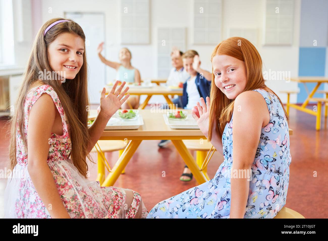 Portrait of smiling female friends waving while sitting in school cafeteria Stock Photo