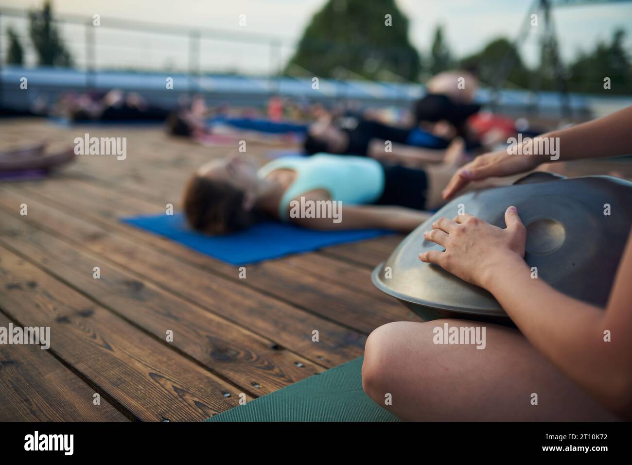 Group of young people practicing yoga lesson lying in Dead Body or Corpse pose outdoor, Shavasana exercise. Hands with drum on a foreground Stock Photo
