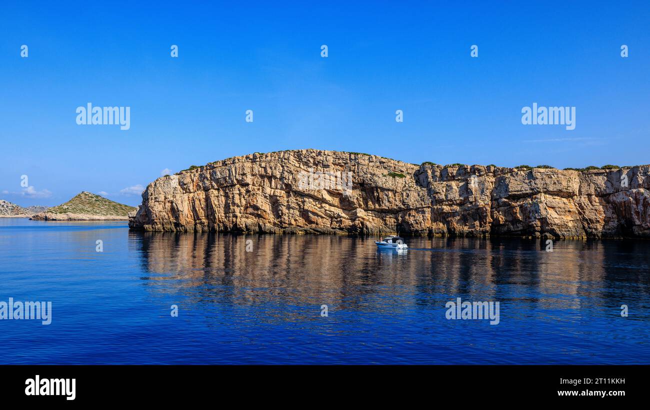 a small boat approaches the sheer limestone cliff of one of the kornati islands reflected in the calm blue sea Stock Photo