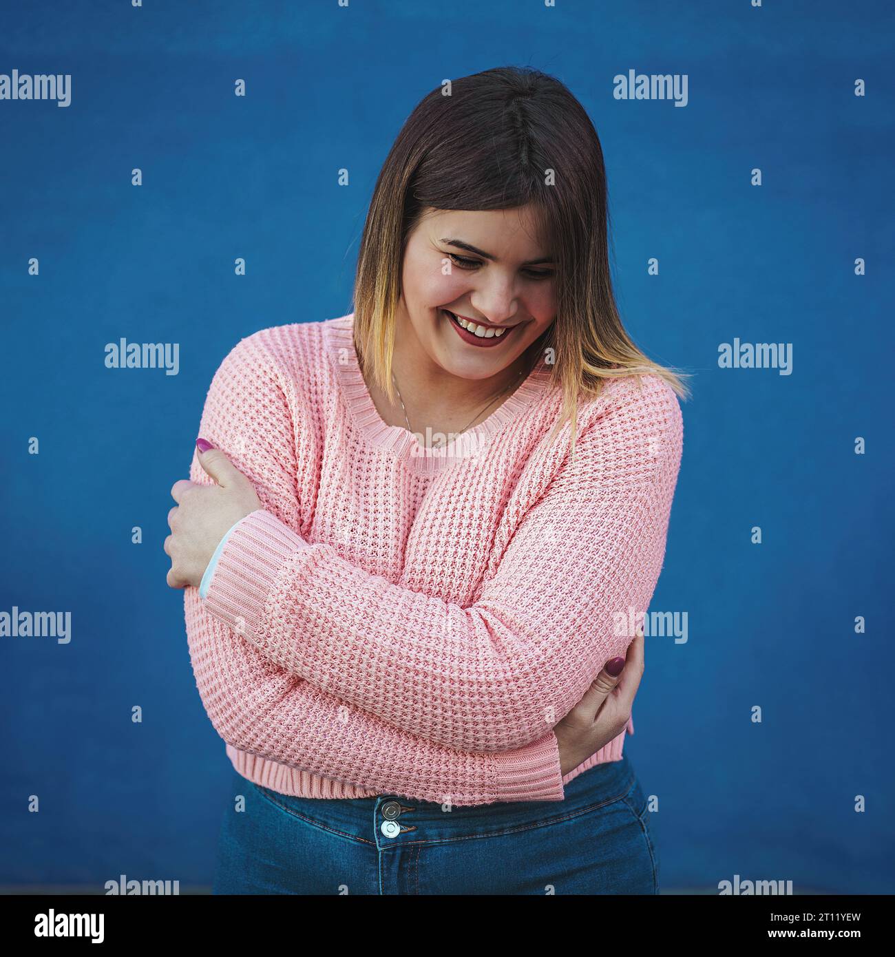 A 20-something curvy caucasian woman with straight hair, in a pink cotton sweater and jeans, hugs herself outdoors, radiating body positivity. Backdro Stock Photo