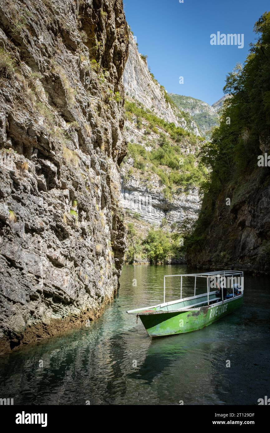 A long thin tour boat moored by a steep rock face at River Shala resort in Lake Komani, Albania Stock Photo