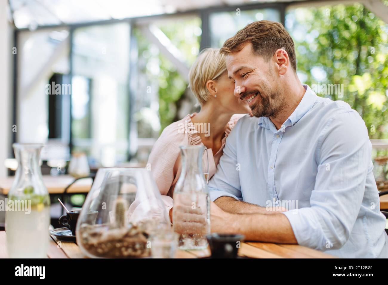 Portrait of beautiful couple in restaurant, on romantic date. Wife whispers into his husband's ear, having romantic moment at restaurant patio. Stock Photo
