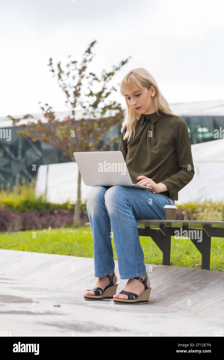 Caucasian blonde girl entrepreneur business park sending an email with laptop. Modern polygon among nature Stock Photo