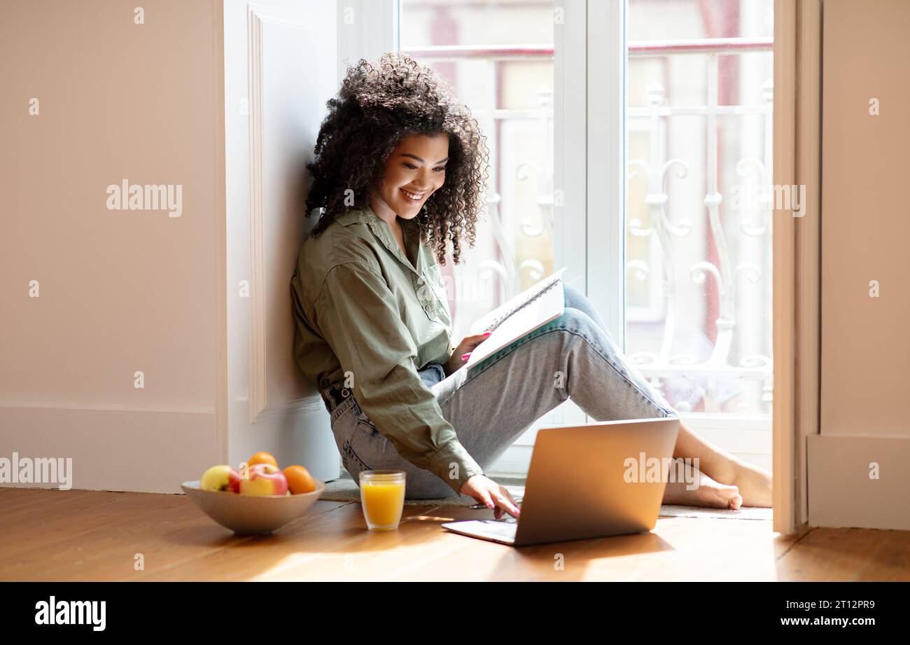 Young black lady using laptop holding notebook for notes indoor Stock Photo