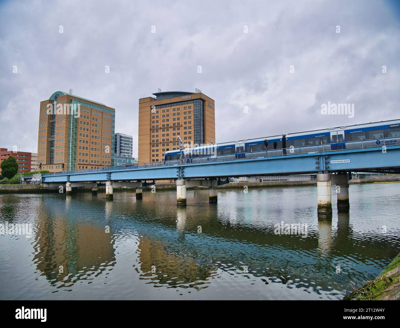 A local commuter train crosses the River Lagan near Lanyon Place Station in Belfast, Northern Ireland, UK. The BT Building and Hilton Hotel appear Stock Photo