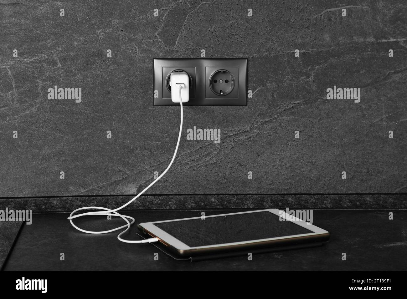 Tablet charging from electric socket on dark grey table indoors Stock Photo