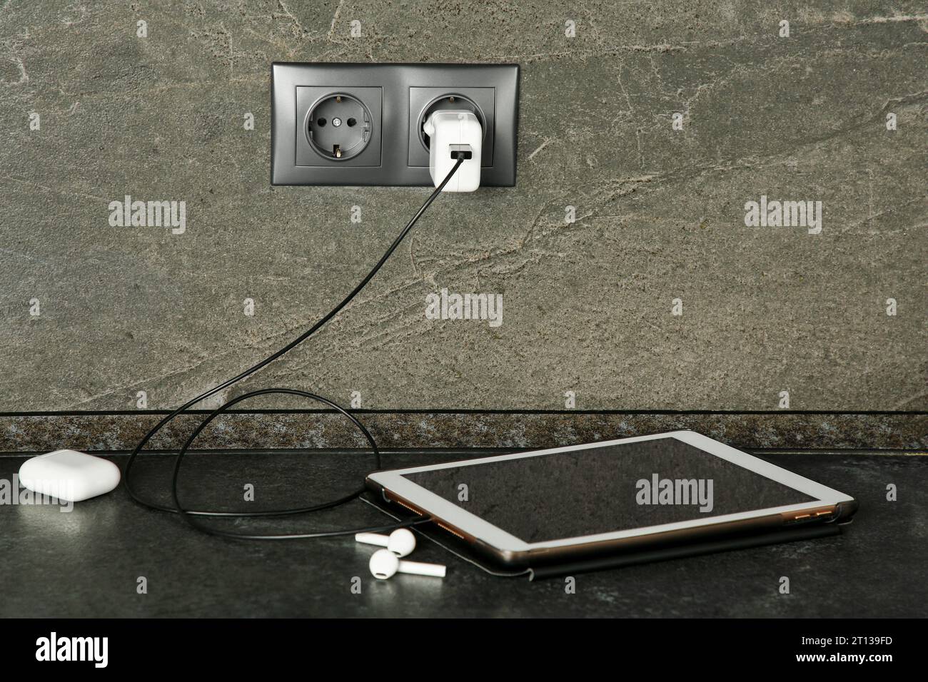 Tablet charging from electric socket on dark grey table indoors Stock Photo