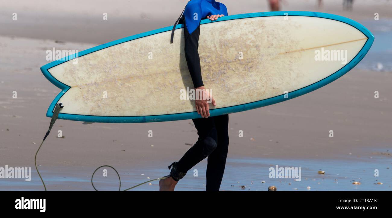 Side view of a surfer wearing a black and blue wetsuit carrying his surfboard into the ocean. Stock Photo