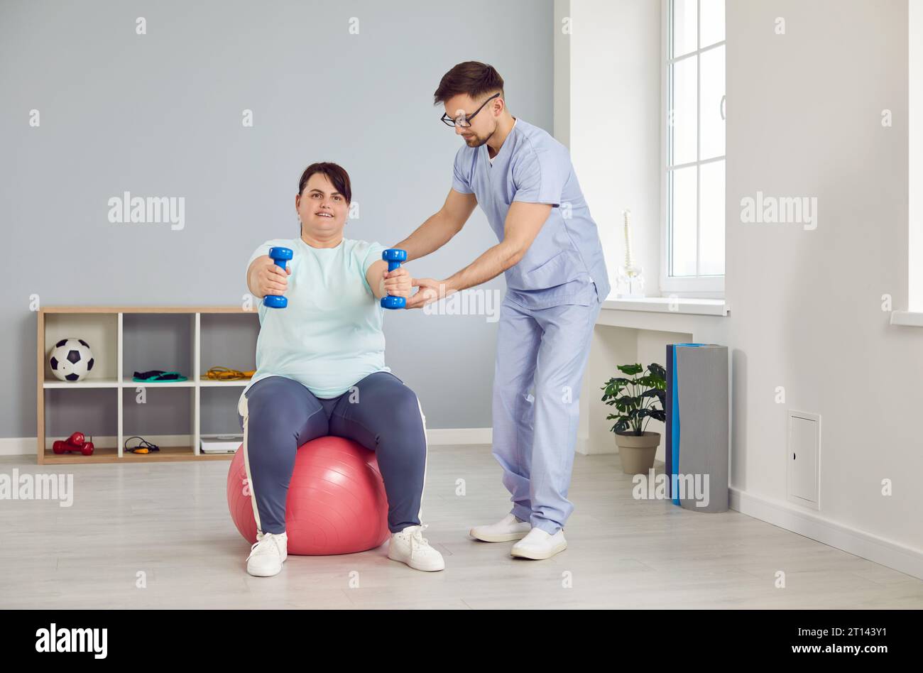 Fat young woman doing sport exercise using dumbbells with support from a male nurse. Stock Photo