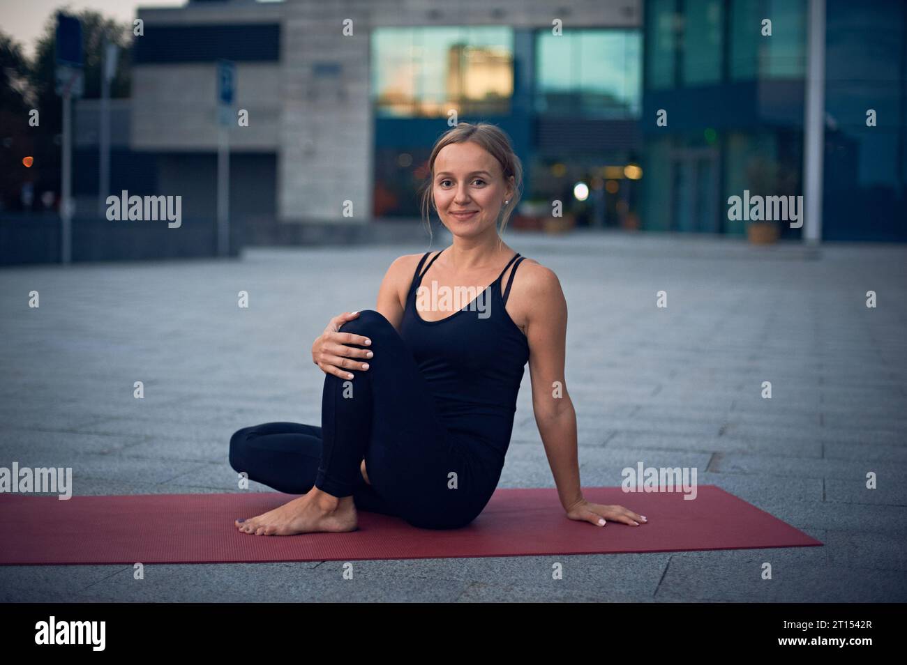 Beautiful young woman prepares to practice yoga asana Marichiasana 4 - sage pose outdoors against the background of a modern city. Stock Photo