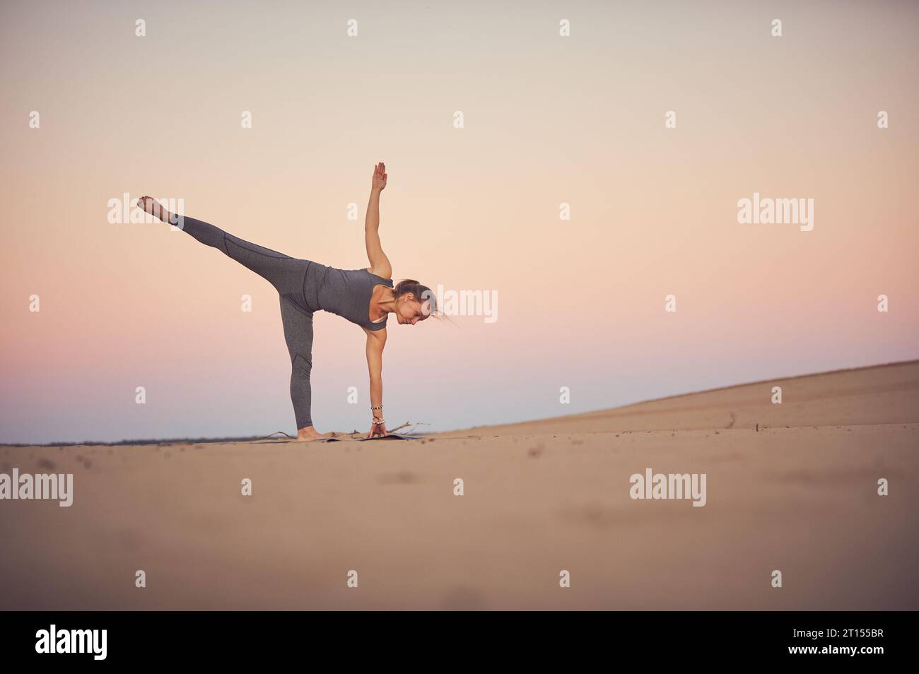 Beautiful young woman practices yoga asana Ardha Chandrasana - Half Moon pose in the desert at sunset. Stock Photo