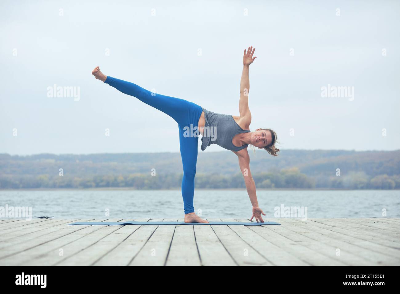 Beautiful young woman practices yoga asana Ardha Chandrasana - Half Moon pose on the wooden deck near the lake. Stock Photo