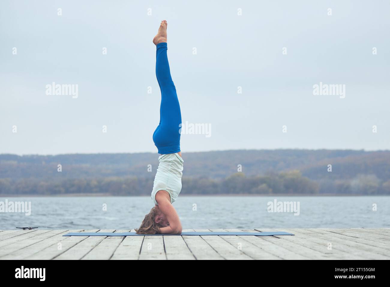 Beautiful young woman practices yoga asana Shirshasana - Headstand pose on the wooden deck near the lake. Stock Photo