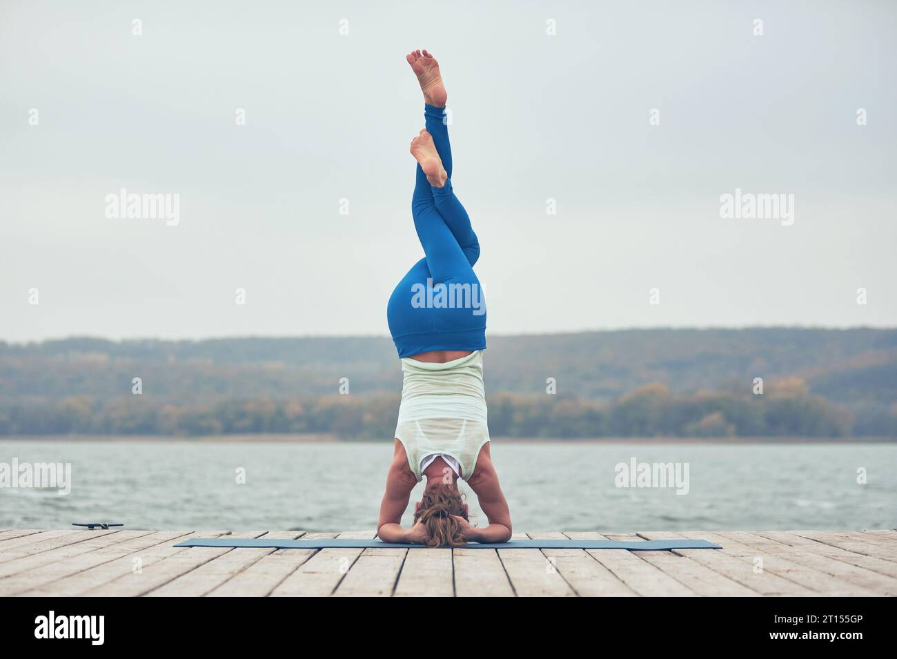 Beautiful young woman practices yoga asana Shirshasana - Headstand pose on the wooden deck near the lake. Stock Photo