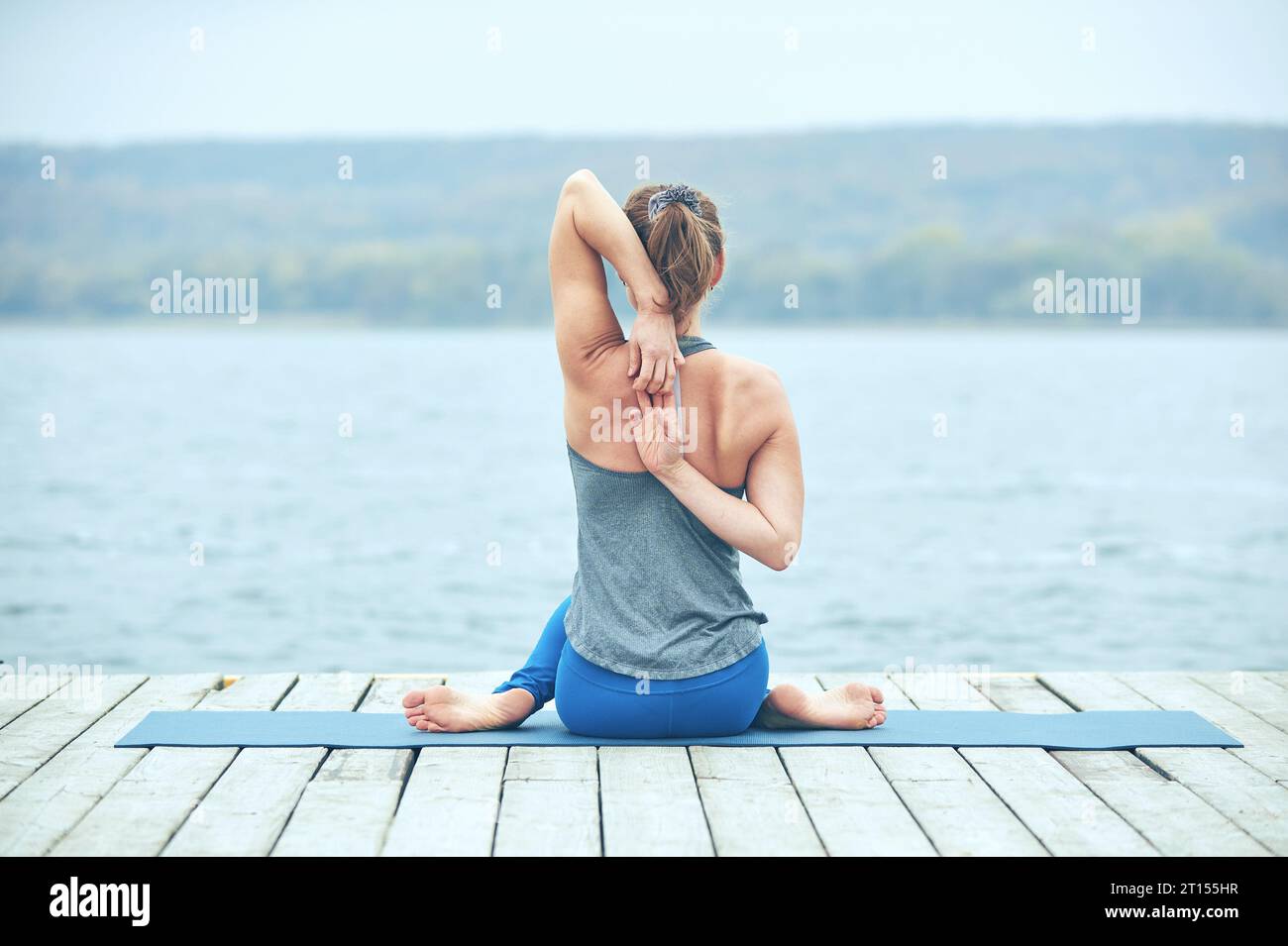 Beautiful young woman practices yoga asana Gomukhasana - Cow face pose on the wooden deck near the lake. Stock Photo