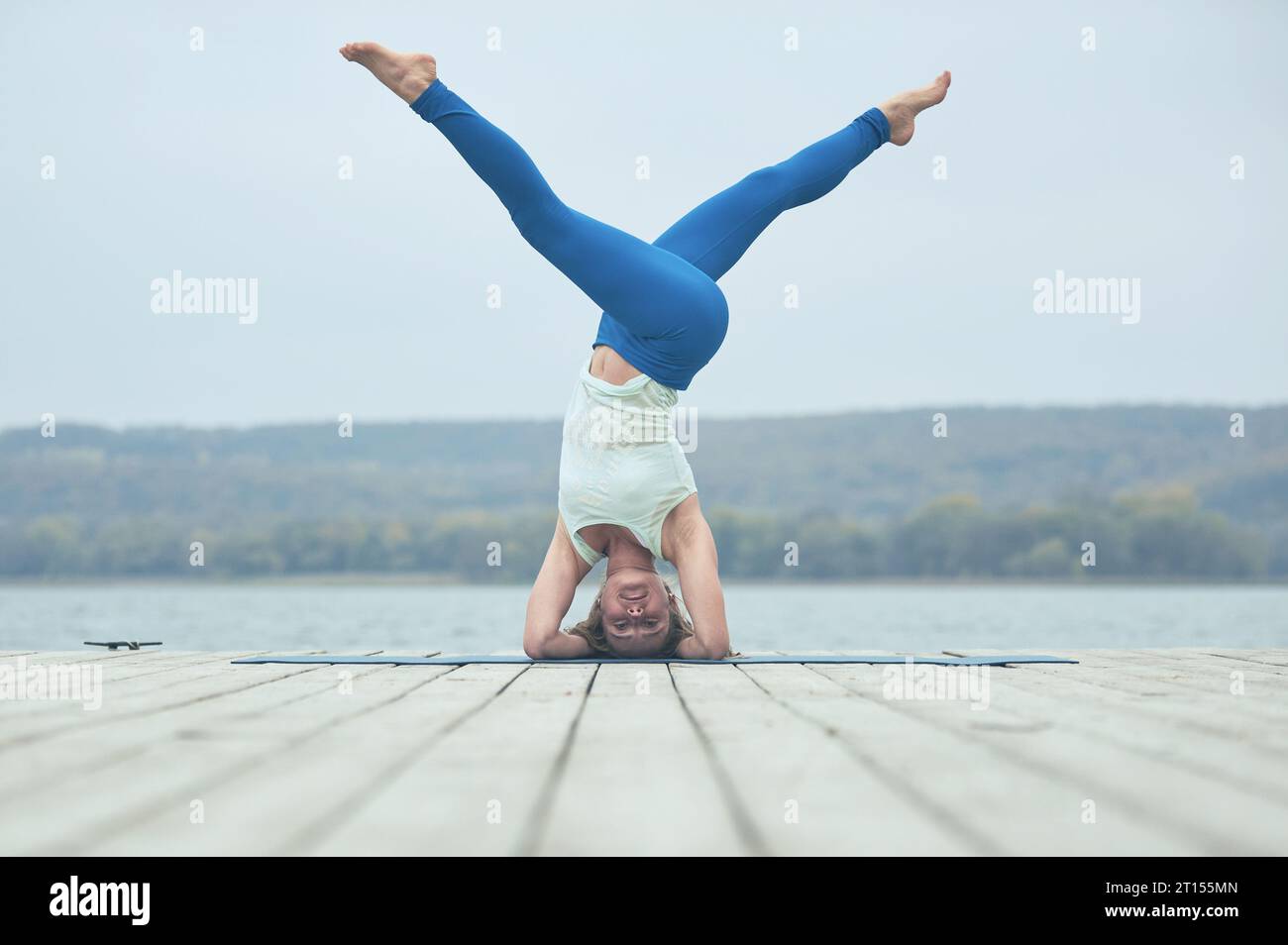 Beautiful young woman practices yoga asana Parshva Salamba Shirshasana - Headstand pose on the wooden deck near the lake. Stock Photo