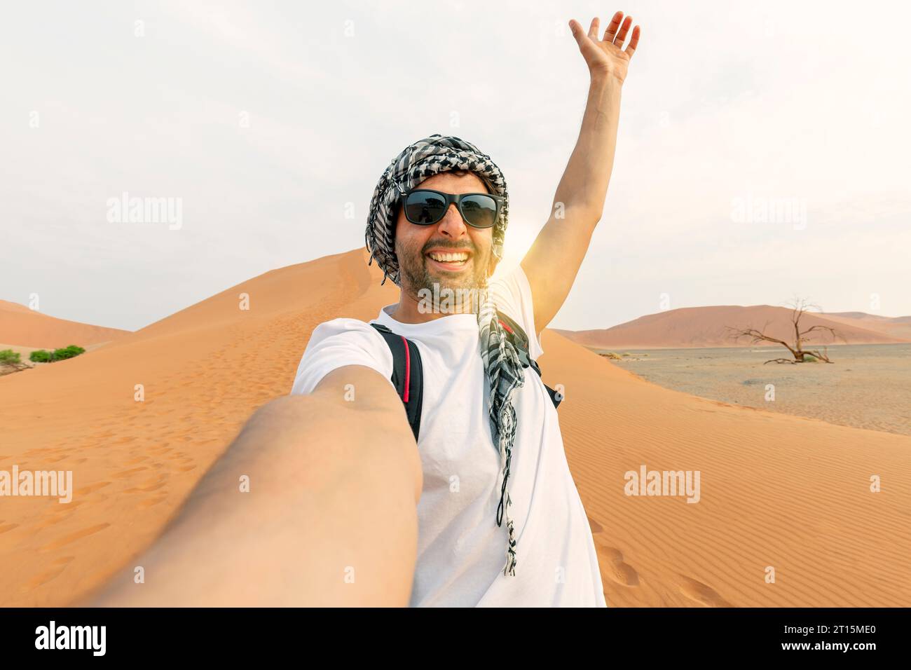 young man in white tshirt in desert, treveling in UAE on safari, wearing hat and backpack, exploring nature of sandy beauty. making selfie in the back Stock Photo