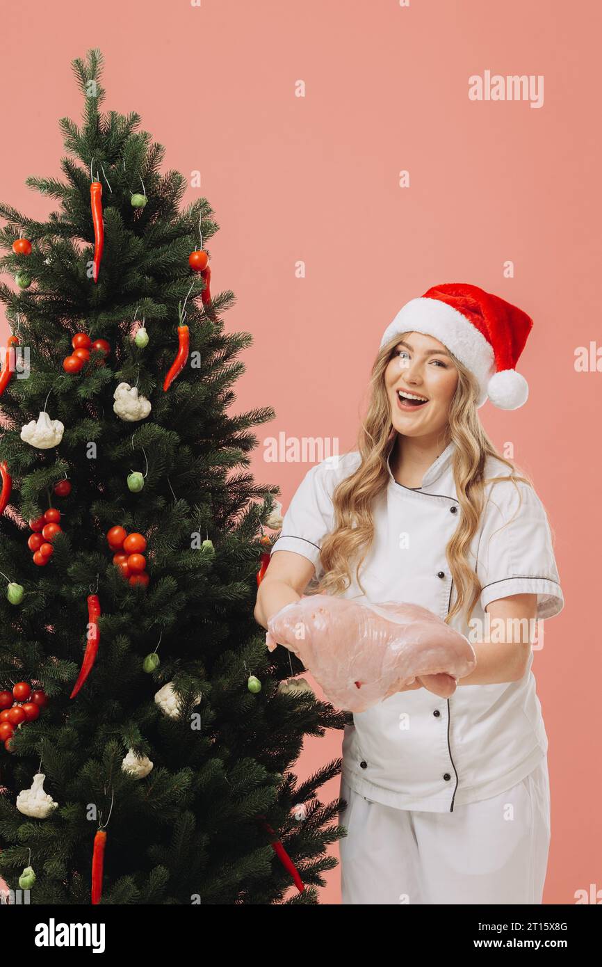 Concept of cooking and Christmas holidays. A beautiful blonde cook in a Santa hat poses against the background of a Christmas tree. Stock Photo