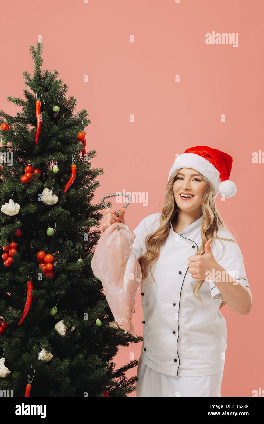 Concept of cooking and Christmas holidays. A beautiful blonde cook in a Santa hat poses against the background of a Christmas tree. Stock Photo