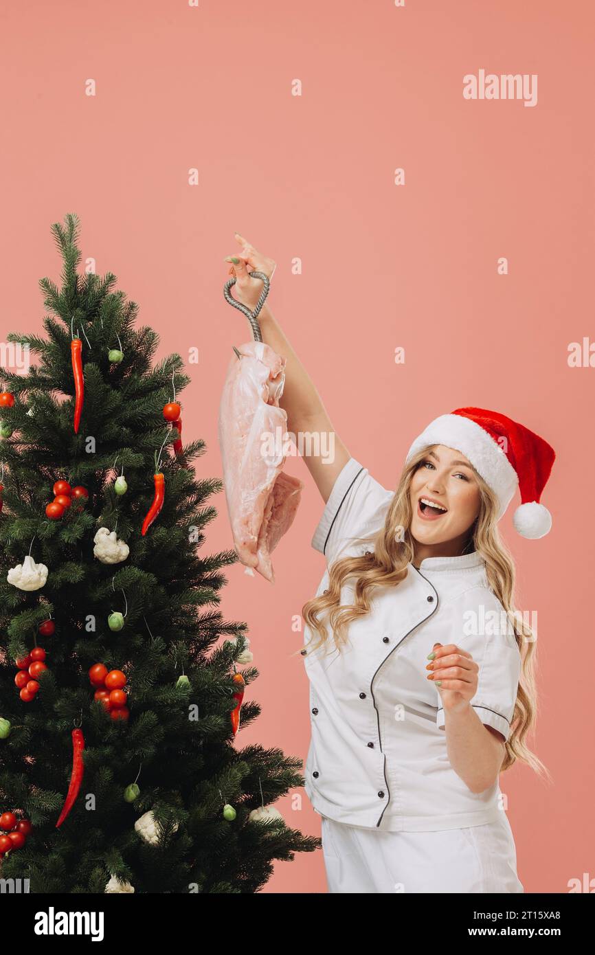 Concept of cooking and Christmas holidays. A beautiful blonde cook in a Santa hat poses against the background of a Christmas tree. Stock Photo