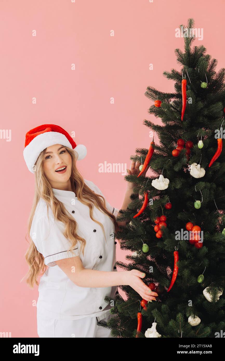 Concept of cooking and Christmas holidays. A beautiful blonde cook in a Santa hat poses against the background of a Christmas tree. Stock Photo