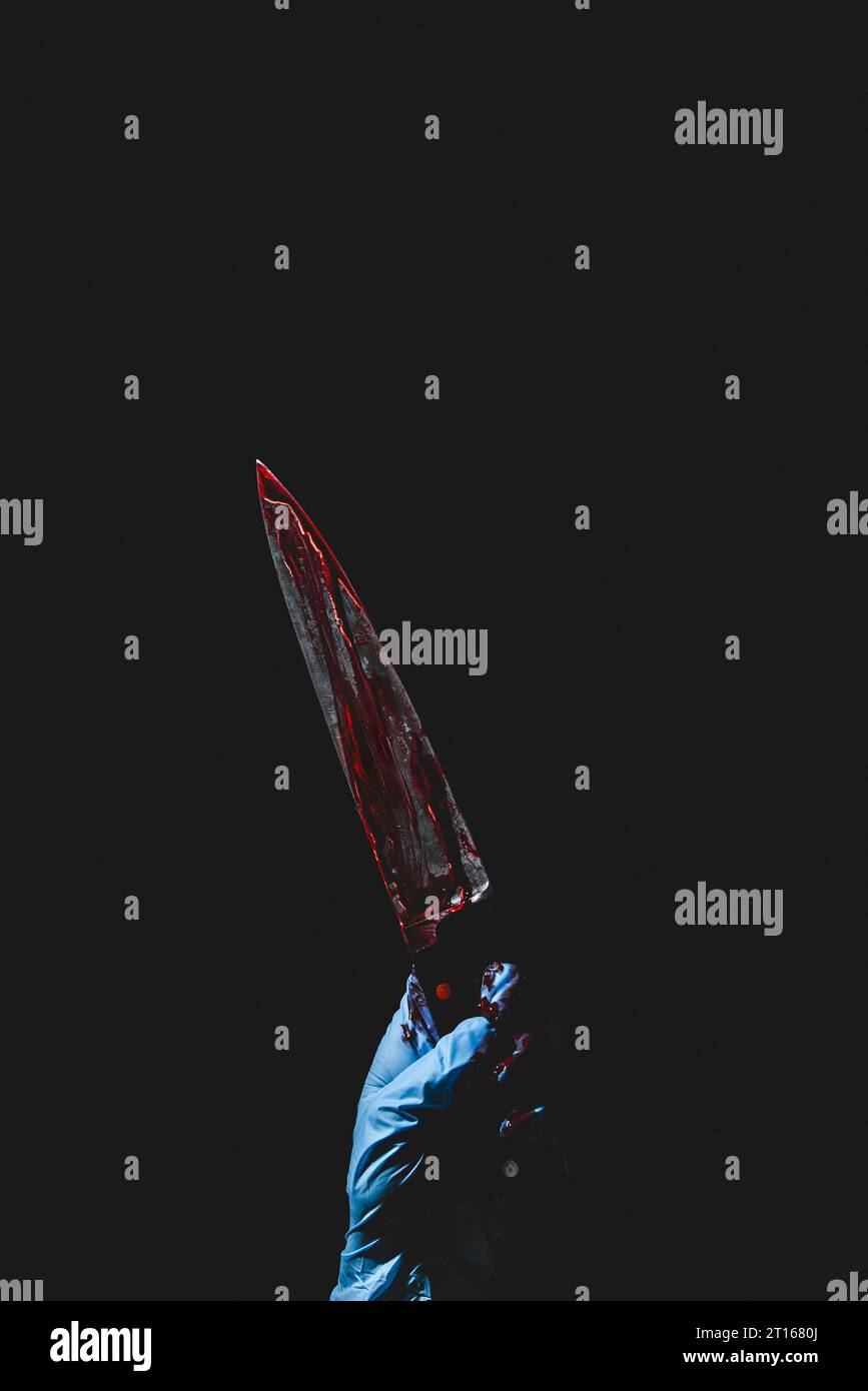 Upward pointing hand in blue glove holds bloodstained knife, dark background, Halloween, copy room Stock Photo