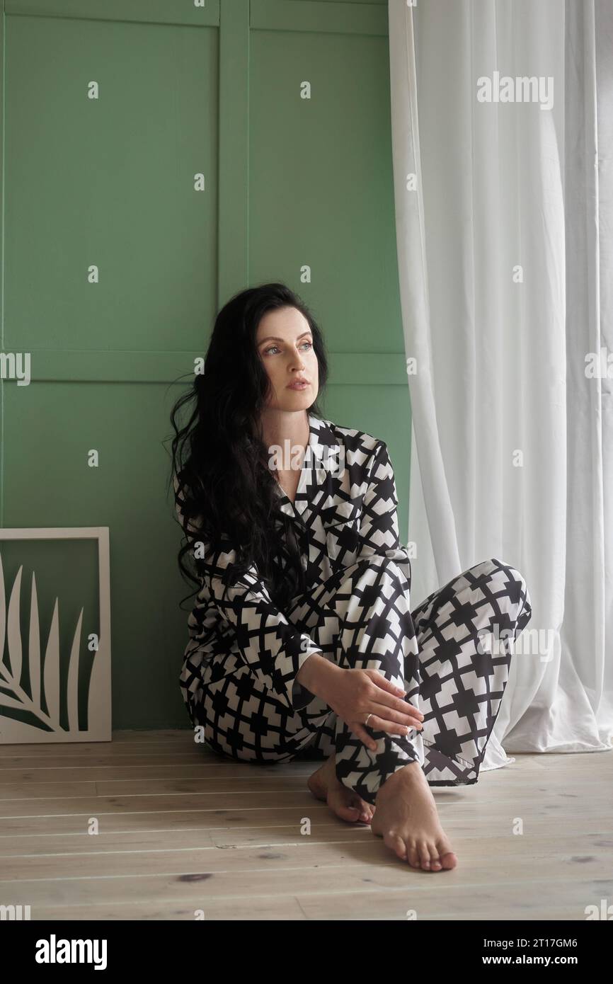 Woman in pajamas is sitting on the floor of the house. Long black brunette hair Stock Photo