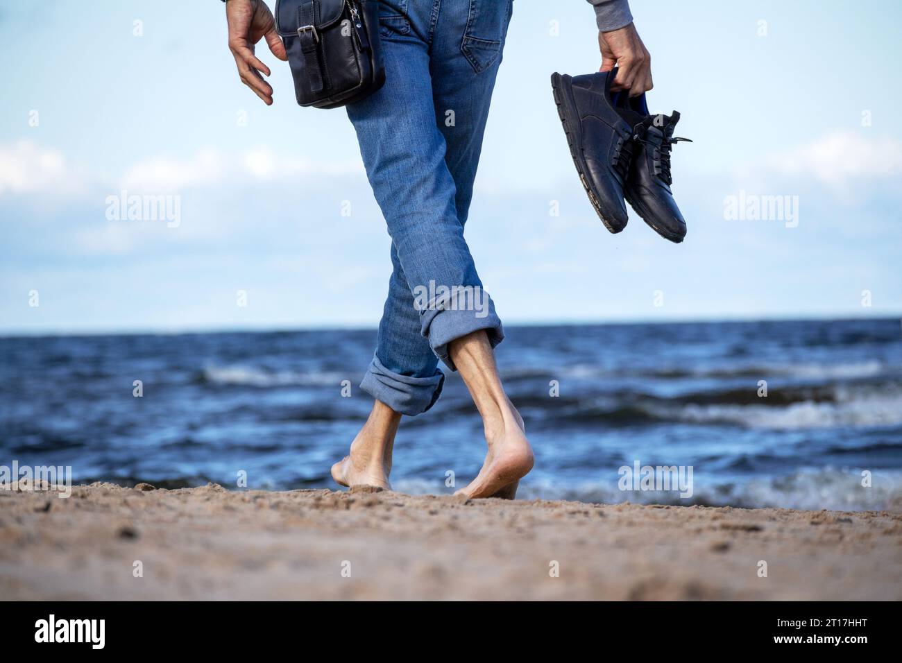 On the seashore of blue color stands a man in denim pants with black shoes in his hands Stock Photo