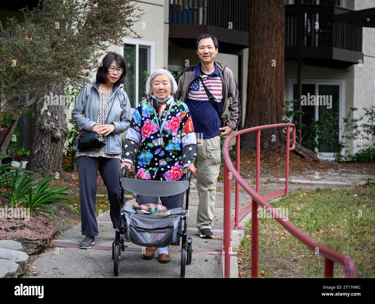 Senior woman with facemask under her chin, walking with family in the courtyard of retirement home. California. Stock Photo