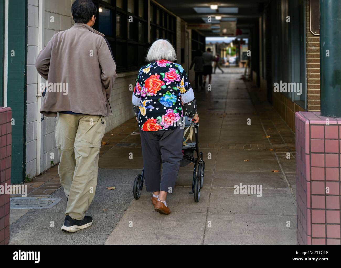 Senior woman walking using a mobility walker in a shopping alleyway, accompanied by her son. California. Stock Photo