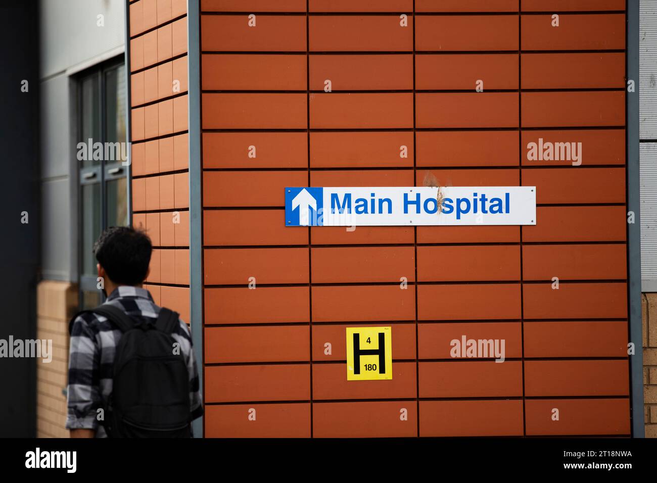 Asian male walking past main entrance to hospital sign with back pack on Stock Photo