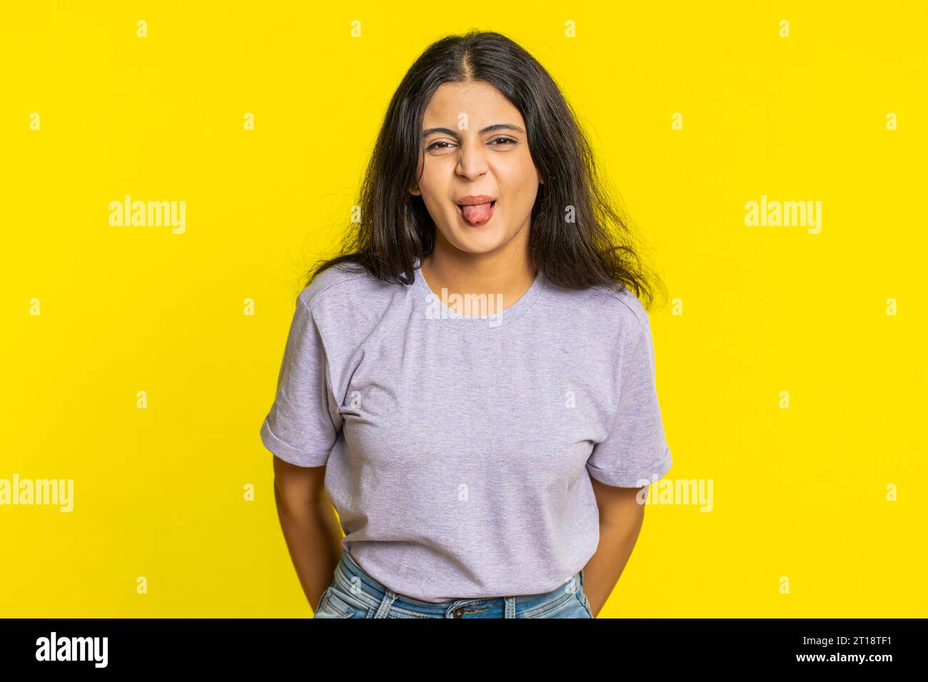 Playful Indian young woman showing tongue making faces at camera, fooling around, joking, aping with silly face, teasing, bullying, abuse. Cheerful funny Arabian girl isolated on yellow background Stock Photo