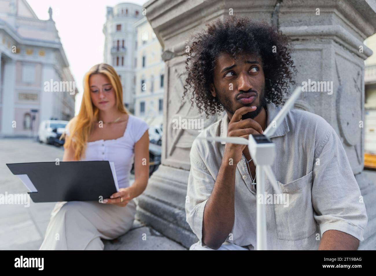 An engineering student is thoughtful as he holds a small wind turbine and his friend goes over her notes Stock Photo