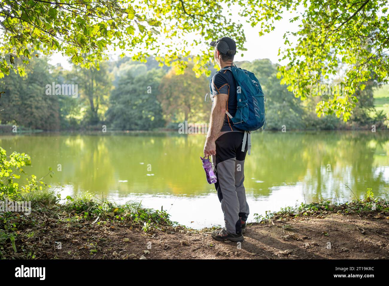 Rear view of a male walker with rucksack on back and water container in hand overlooking a lake on a summer day. Stock Photo