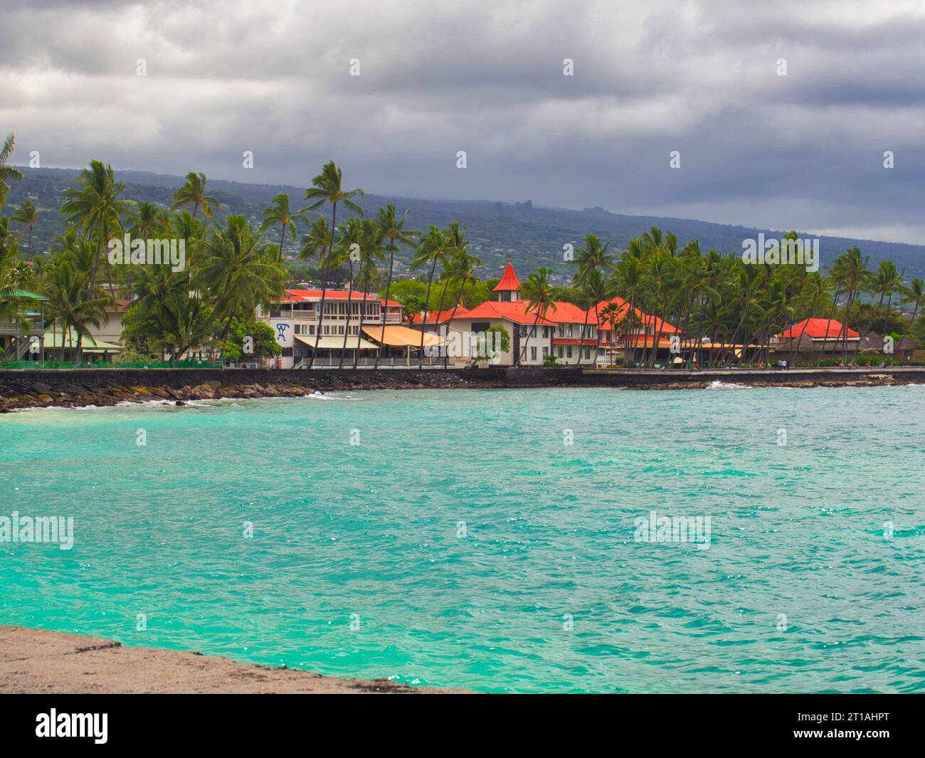 Ocean with very blue water looking over Kailua-Kona downtown with palm trees and red roofs. Storm clouds move in from behind the town. Stock Photo