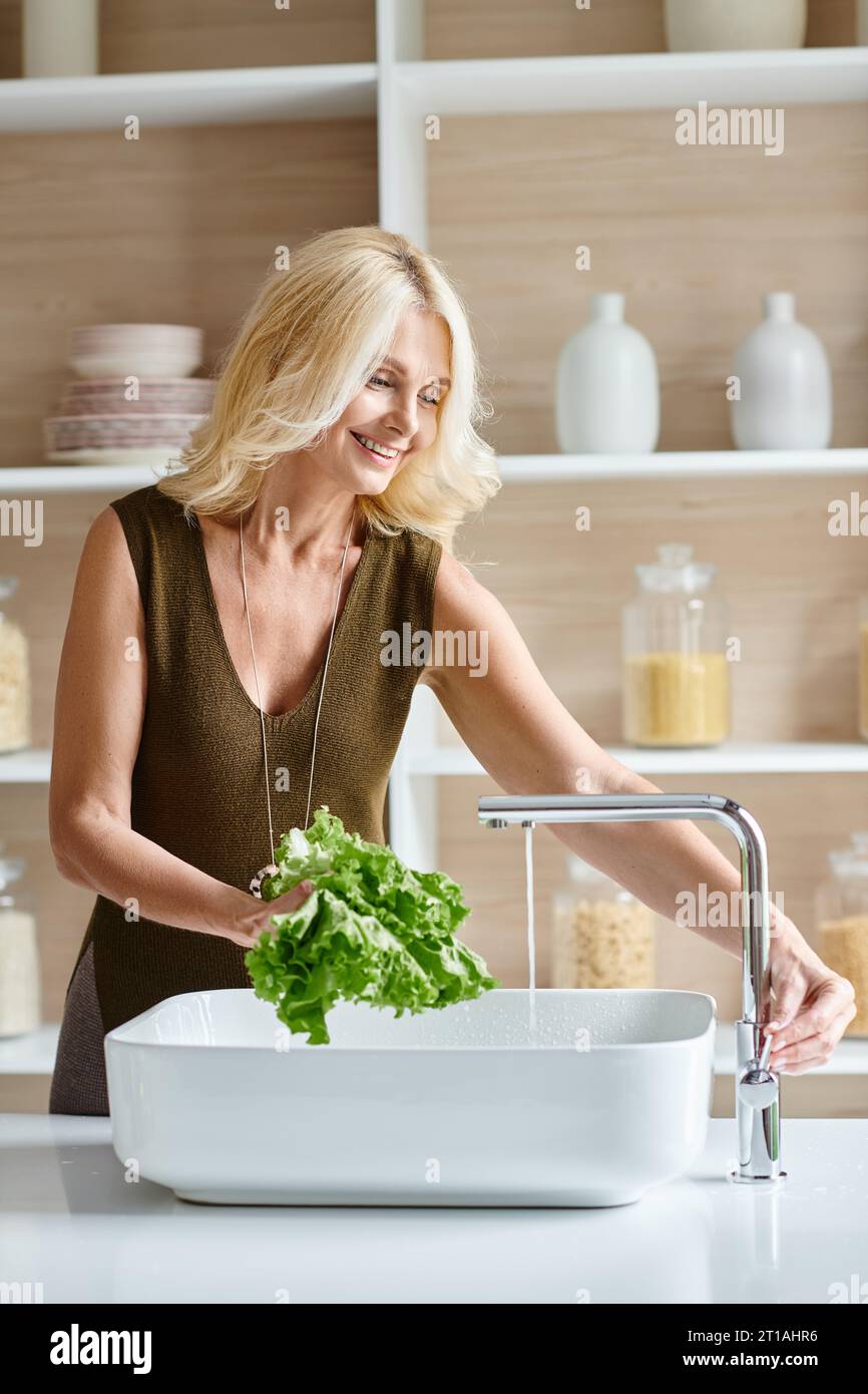 positive middle aged vegetarian woman with blonde hair washing fresh lettuce, vertical shot Stock Photo