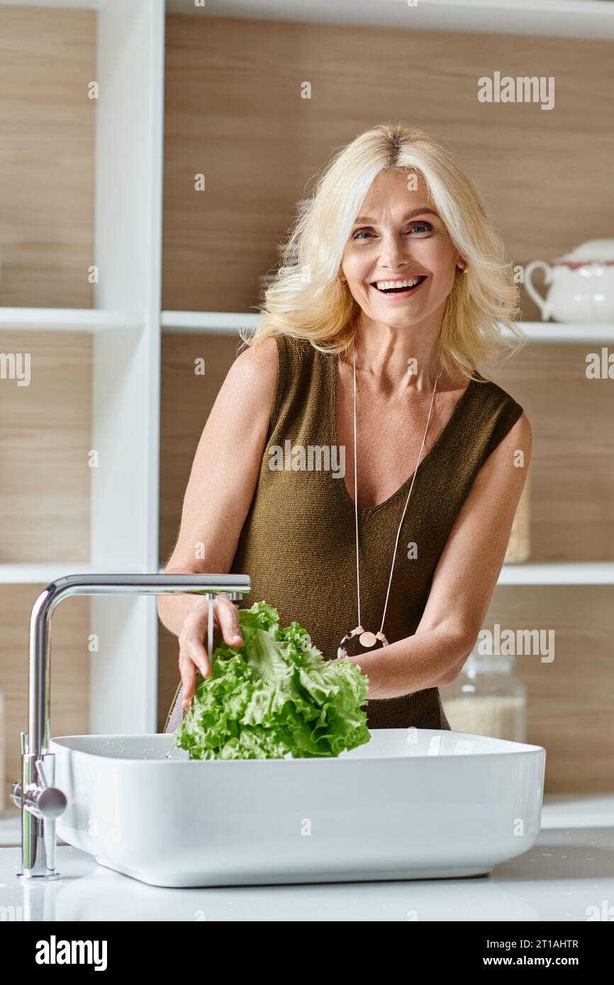 excited middle aged vegetarian woman with blonde hair washing fresh lettuce, vertical shot Stock Photo