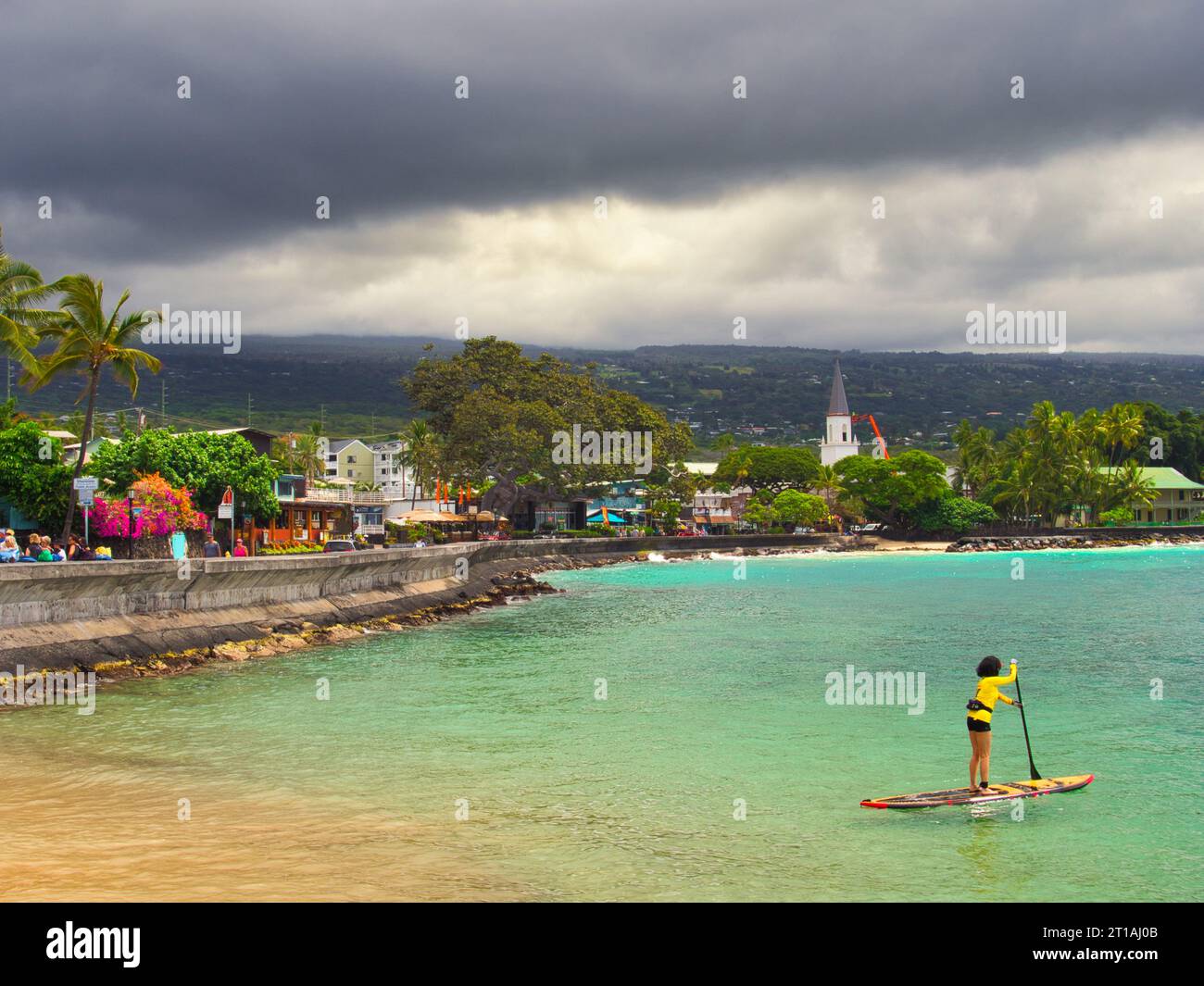 Beach and ocean view of downtown Kailua-Kona, Hawaii on the Big Island with stormy clouds and a paddle boarder in yellow wetsuit on very blue ocean. Stock Photo